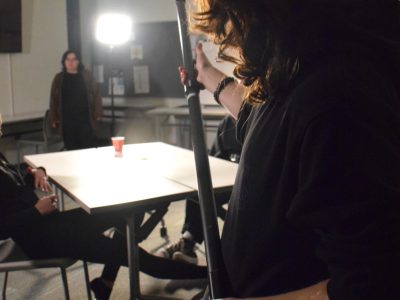 A man in silhouette is holding a boom mic and wearing headphones. He is reaching the boom towards a table where a scene is being shot. It is in a dark classroom setting