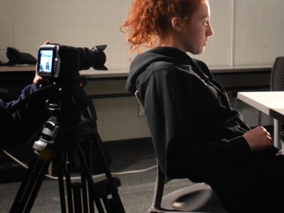 A woman performer with red hair in sitting at a table preforming a scene. Behind her is a camera on a tripod recording the scene.
