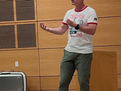 A man in a white t-shirt stands on a stage and gestures as he speaks