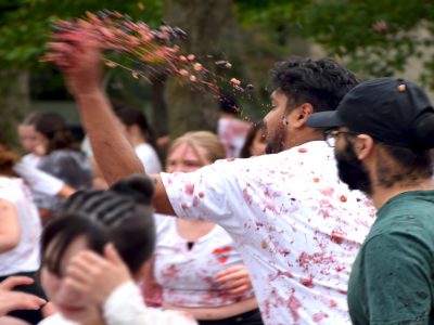 A student in white t-shirt, splattered with red and purple splotches, throws a handful of grapes into the crowd.