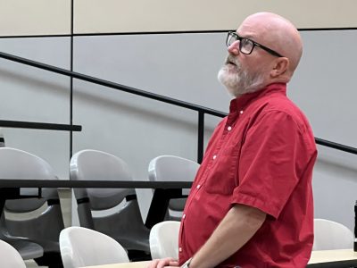 A man in a red shirt stands infront of rows of desks and address an unseen group
