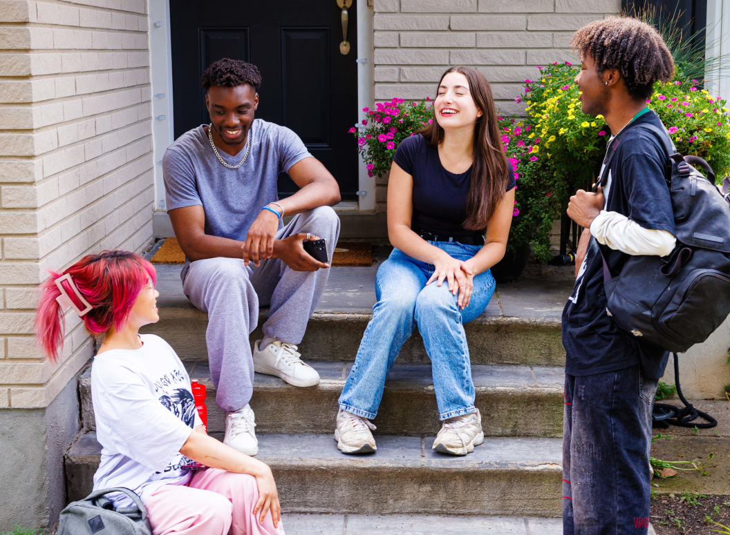 Four students sitting and standing around a front porch. They are speaking and laughing.