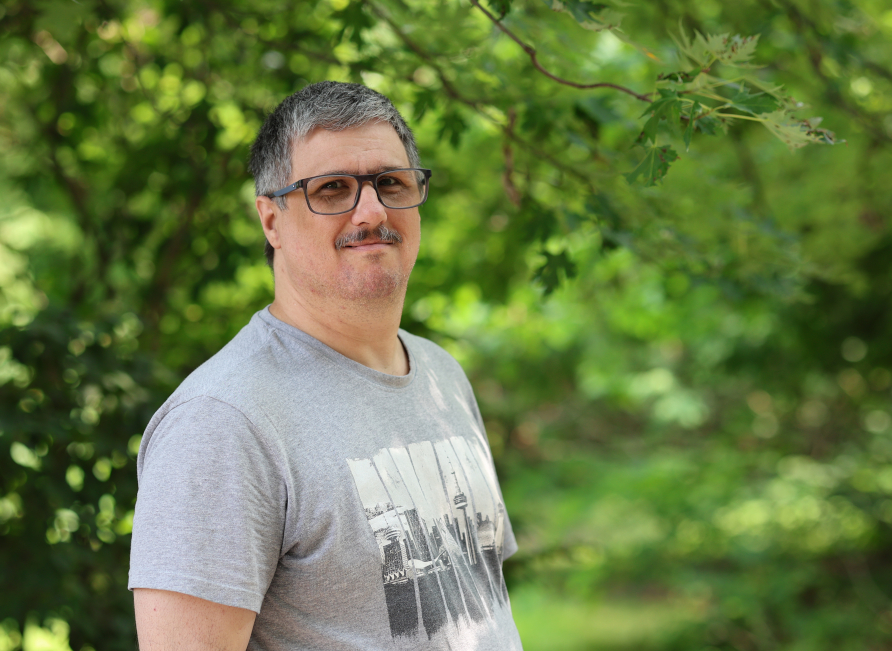 a photo of a man in a gray t-shirt standing in front of a trees and plants.