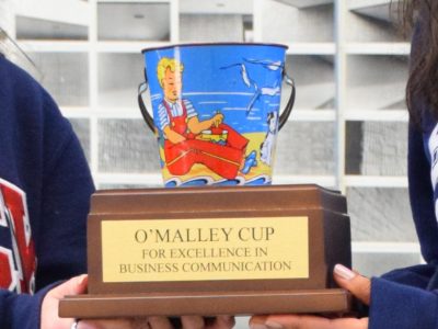 A close up on hands holding a trophy. The trophy is and old, colourful bucket, attached o a base that reads: O'Malley Cup for excellence in business communication