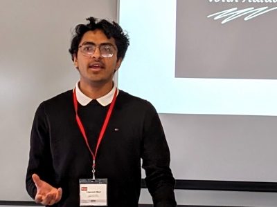 A student in business attire gestures as they give a presentation in front of a projector screen.
