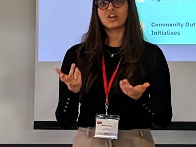 A student in business attire gestures as they give a presentation in front of a projector screen.