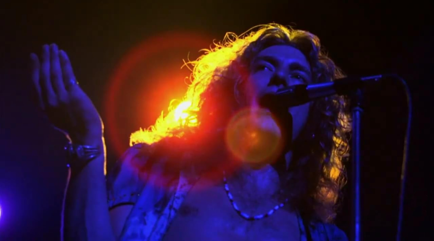 A dark photo of a man with long hair singing. There is a bright lights behind him lighting up the edges of his hair.