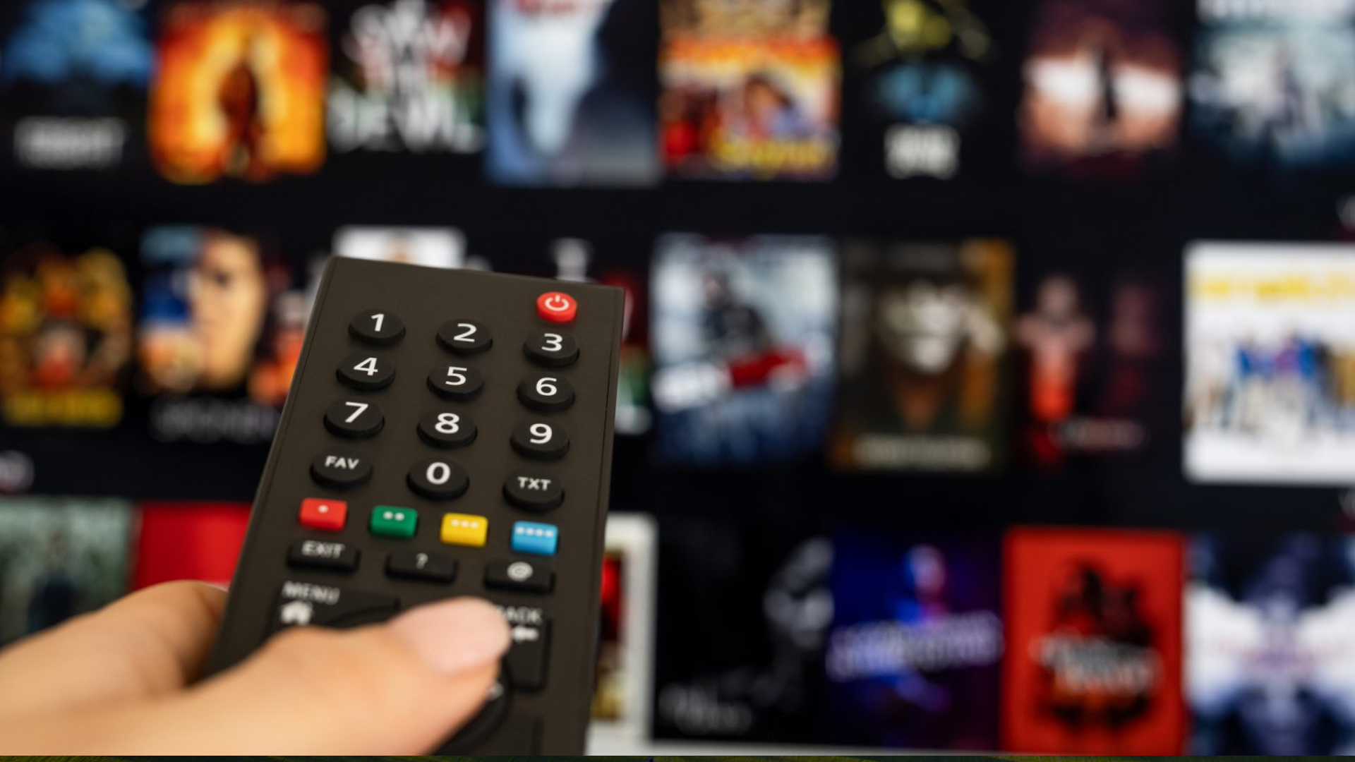 A close-up of a hand holding a remote control in front of a screen showing a list of film choices 