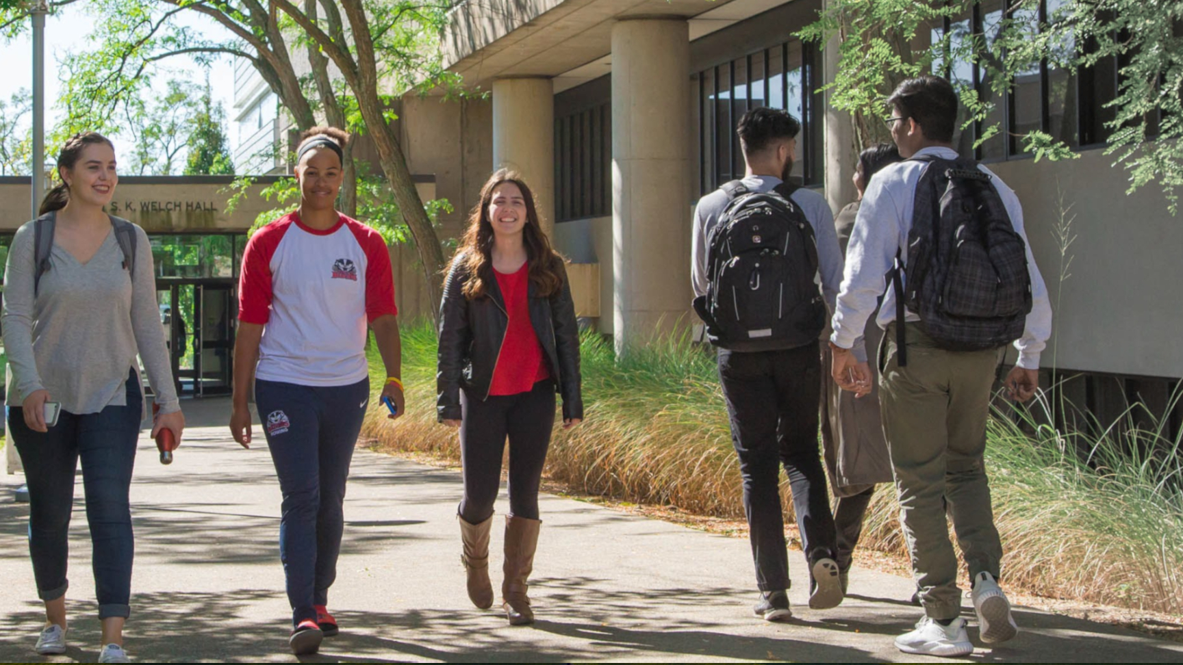 3 students walk along a sidewalk between campus buildings. Two students pass them in the opposite direction