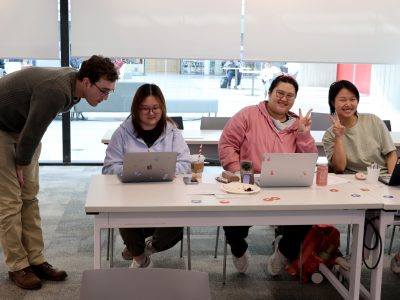 A professor looking over a students work, while two other students pose for the photo