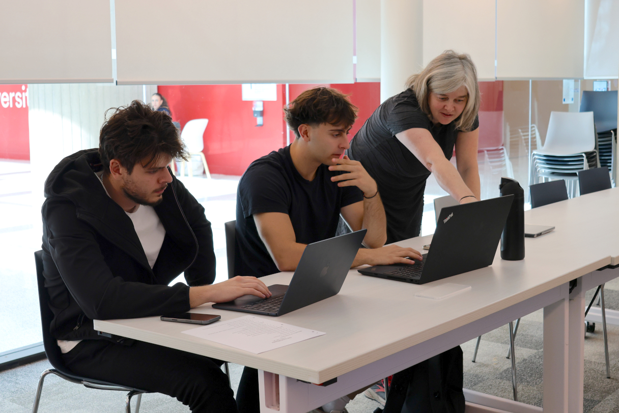 A professor points to a laptop while explaining something to two students