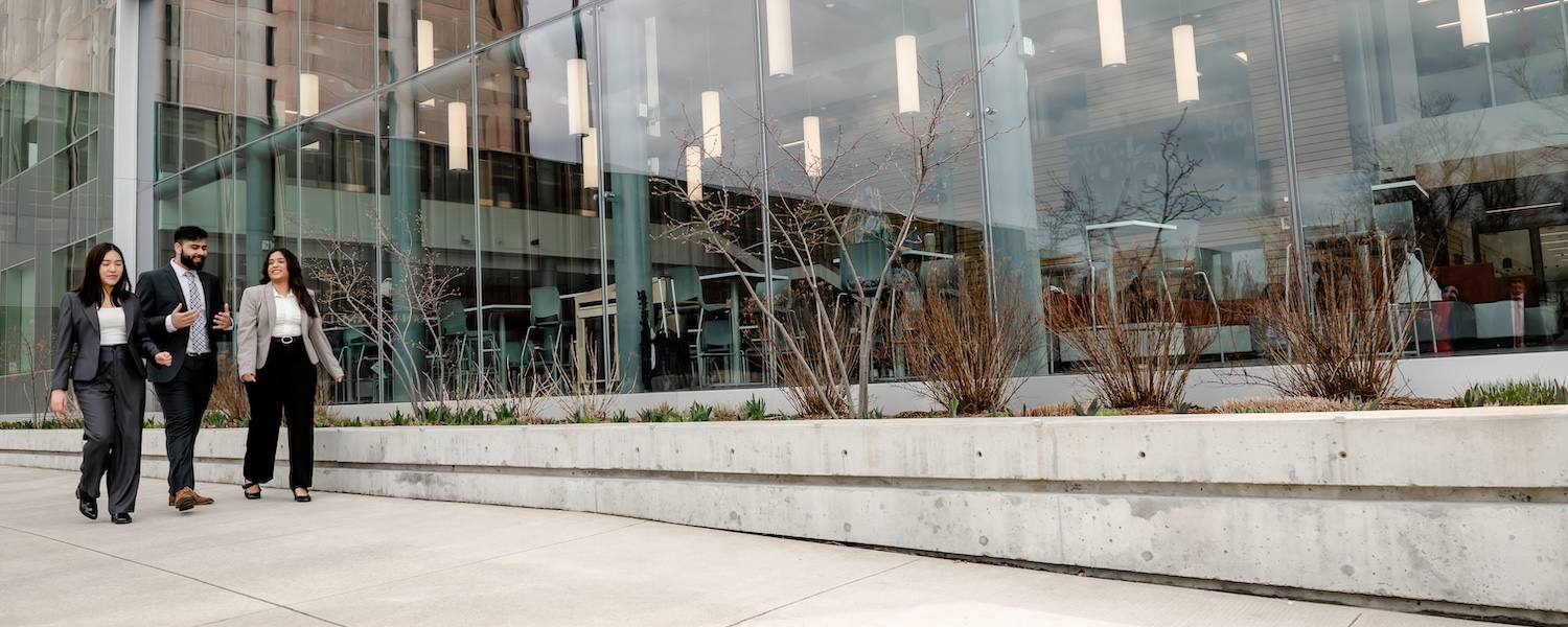 Three students in suits walking outside along a glass building.