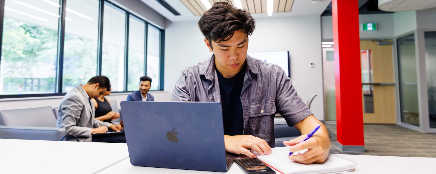 A student sitting at a desk and writing in a notebook with a calculator and laptop next to them and a group of three students visible in the background.