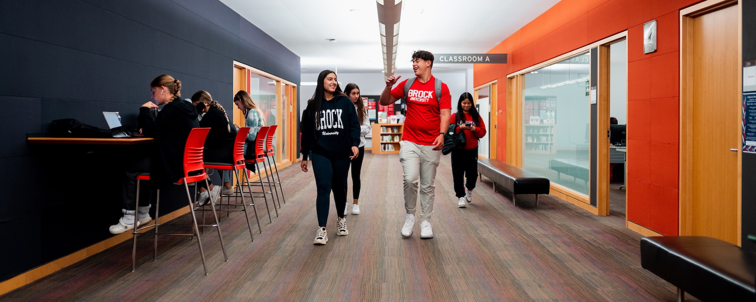 A group of four students wearing Brock University shirts and walking down a hallway with students sitting at a side table on laptops.