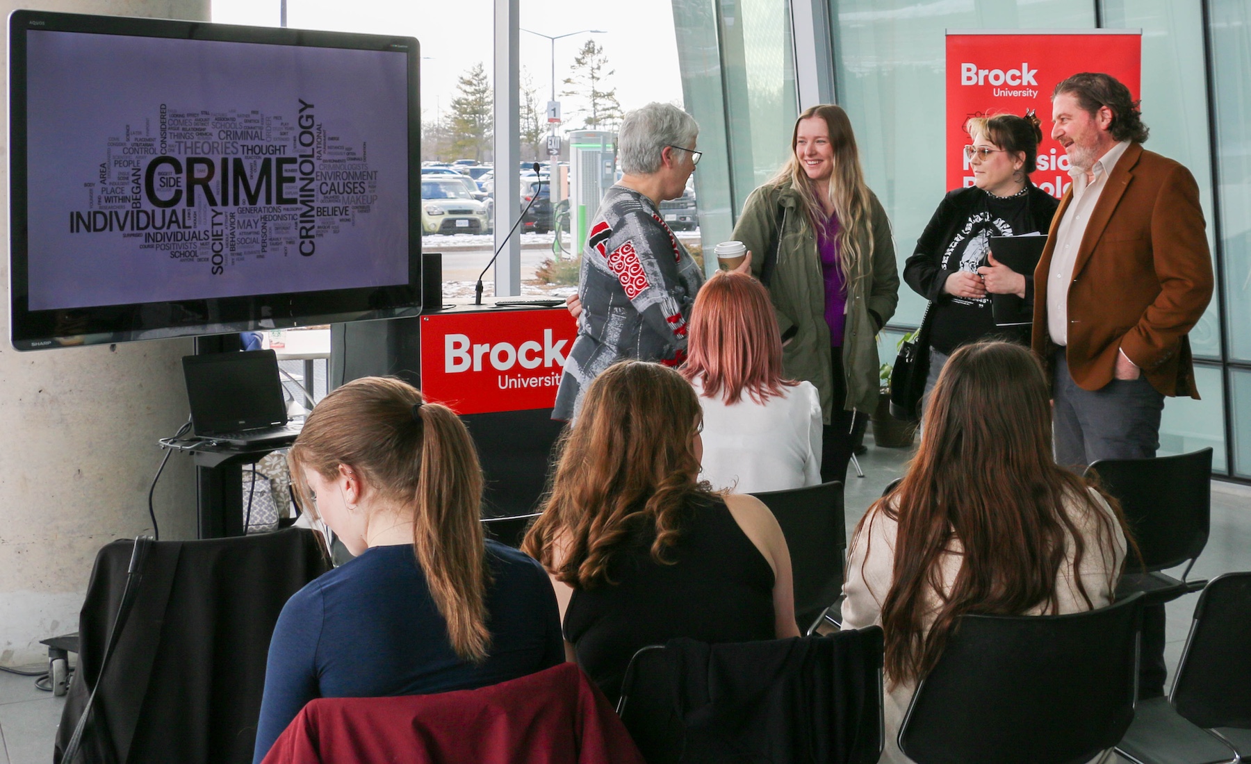 A group of four people talk near a Brock University lectern in front of a seated audience.