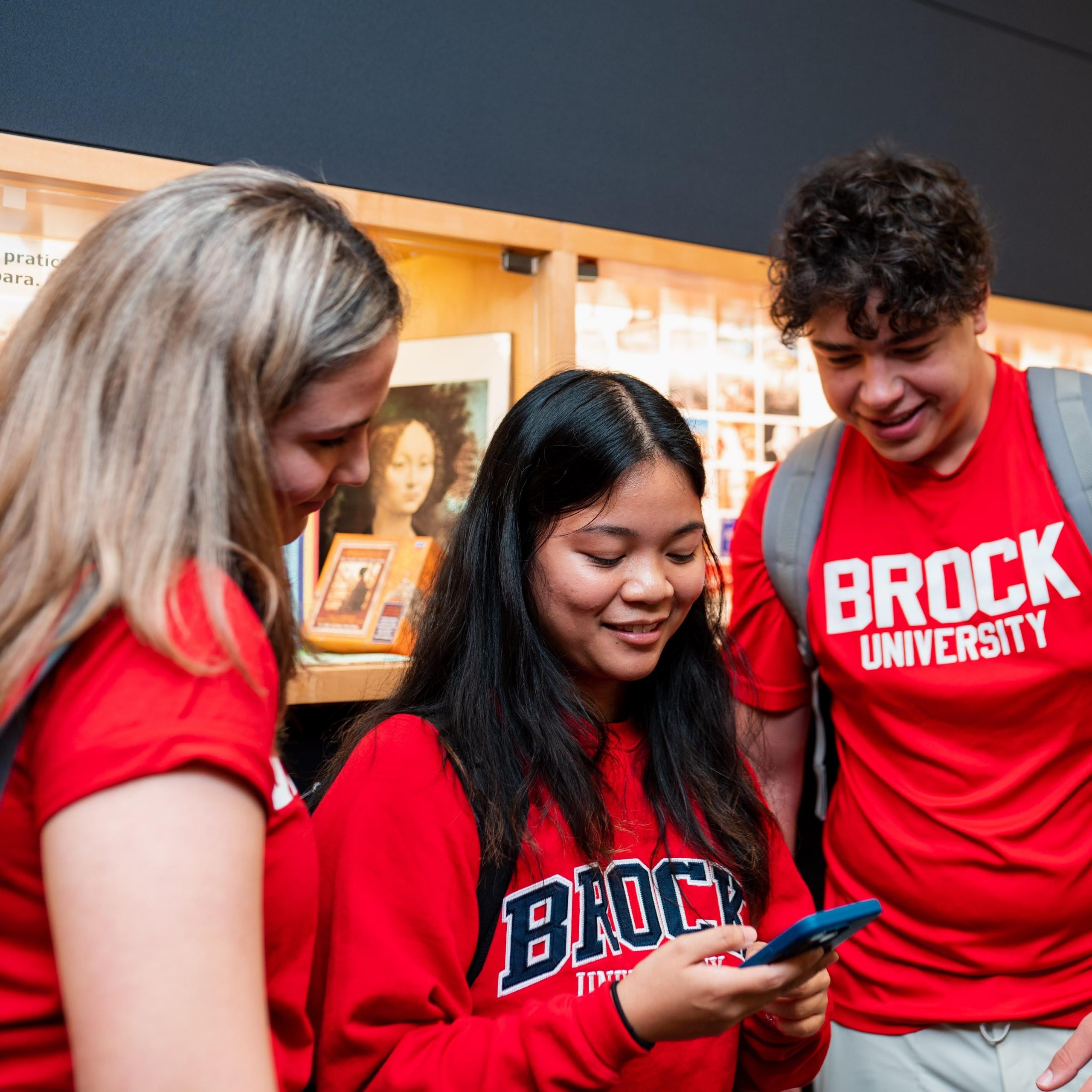 Three students in Brock University shirts looking at a phone together with library displays in the background.