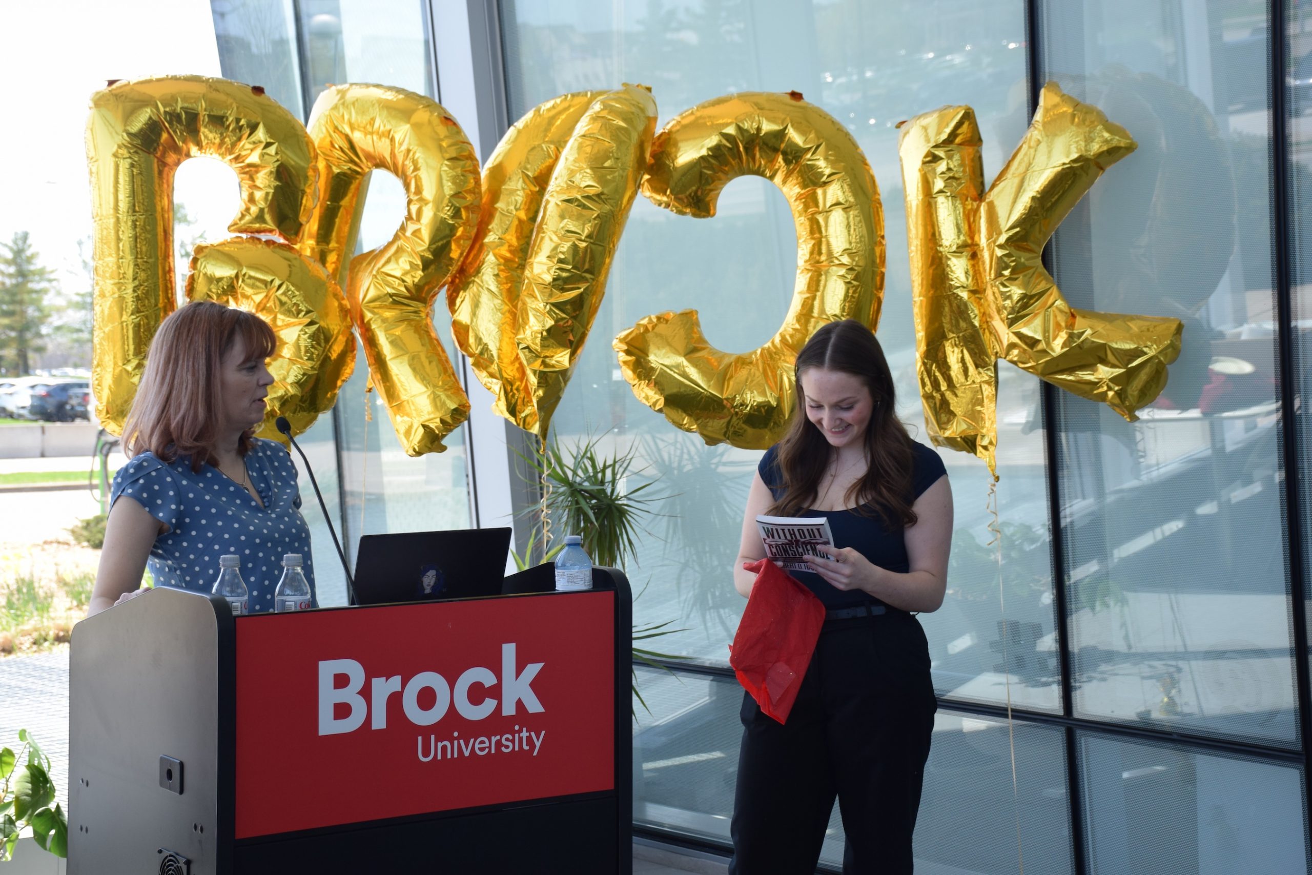 A person smiles down at a book they're holding while a person standing behind a Brock University lectern looks at them and there are balloons that spell Brock in the background.