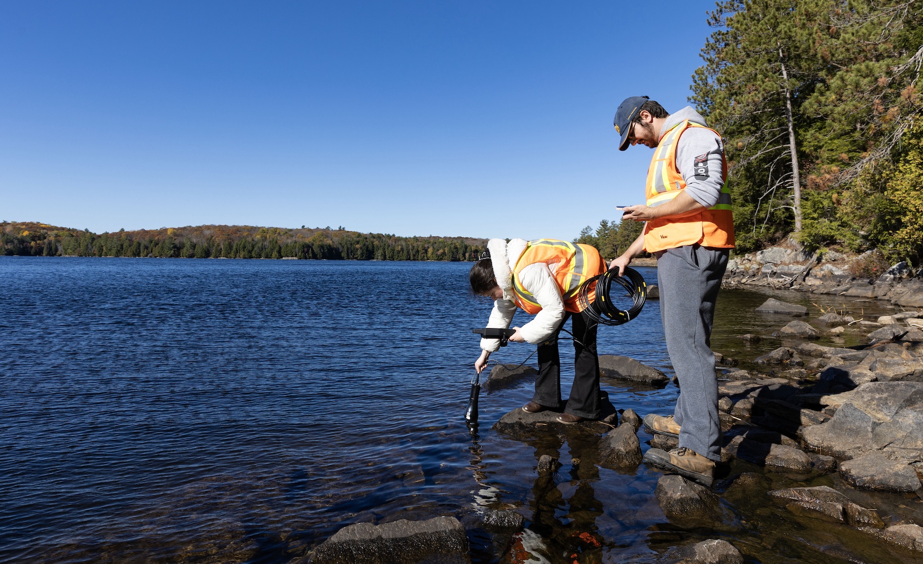 Two people in orange vests standing on rocks in a lake and taking water samples. 