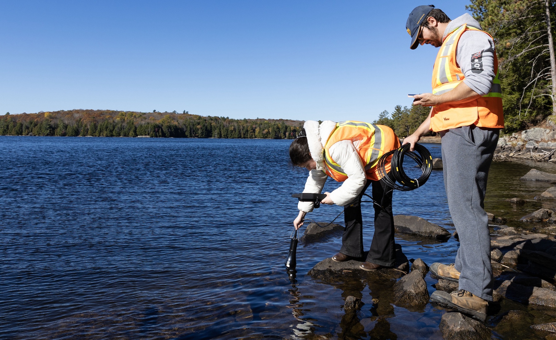 Two people in orange vests standing on rocks in a lake and taking water samples. 