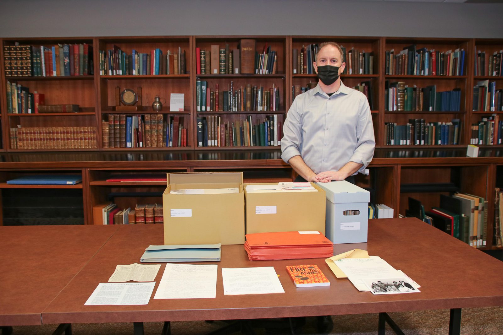 Person standing behind a table with boxes and documents spread out and bookcases in the background.