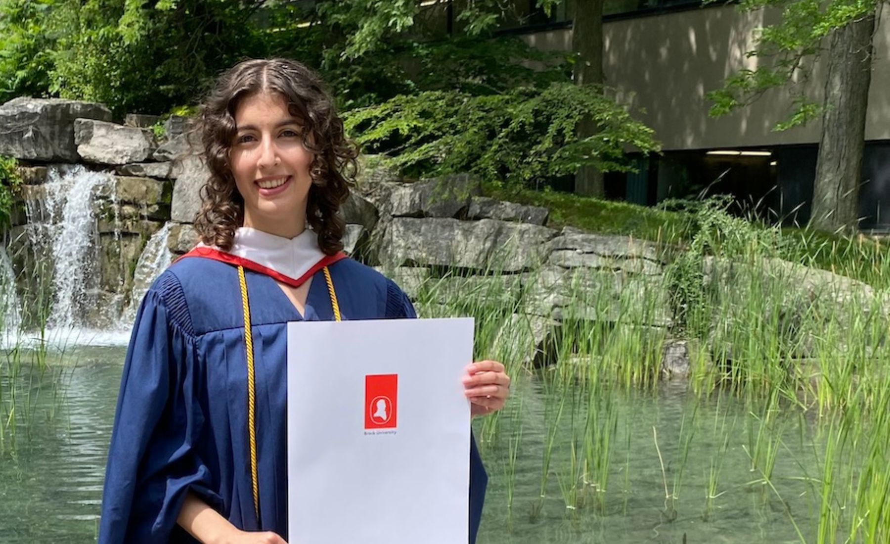 Brontë Slote (BA ’24) standing in Brock University's Pond Inlet wearing a graduation gown and holding up an envelope with the Brock University logo. 