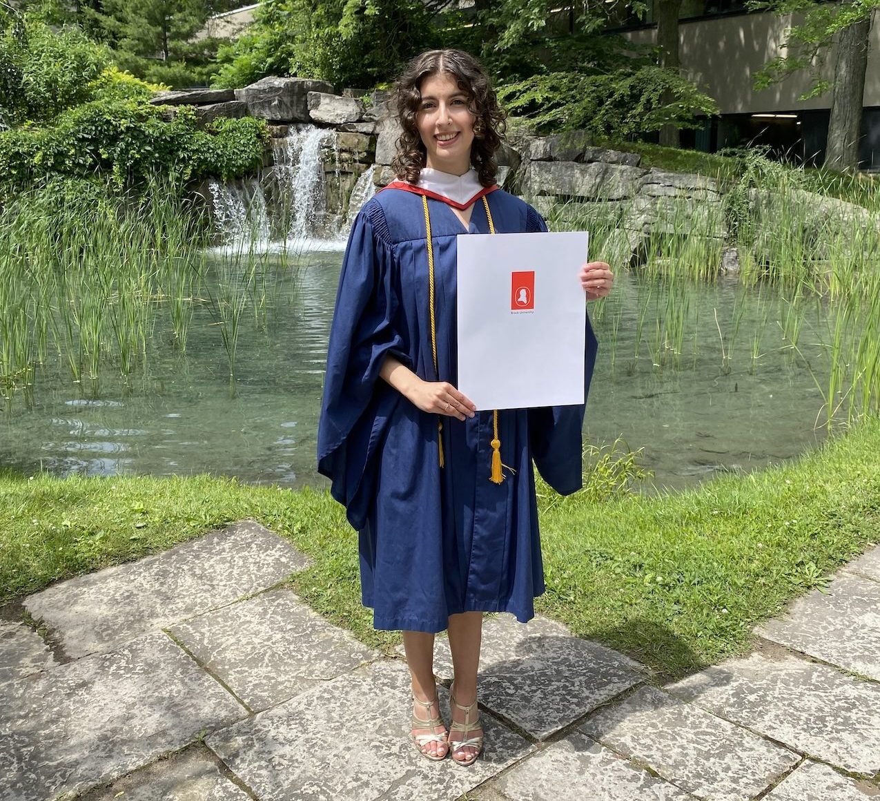 Brontë Slote (BA ’24) standing in Brock University's Pond Inlet wearing a graduation gown and holding up an envelope with the Brock University logo.