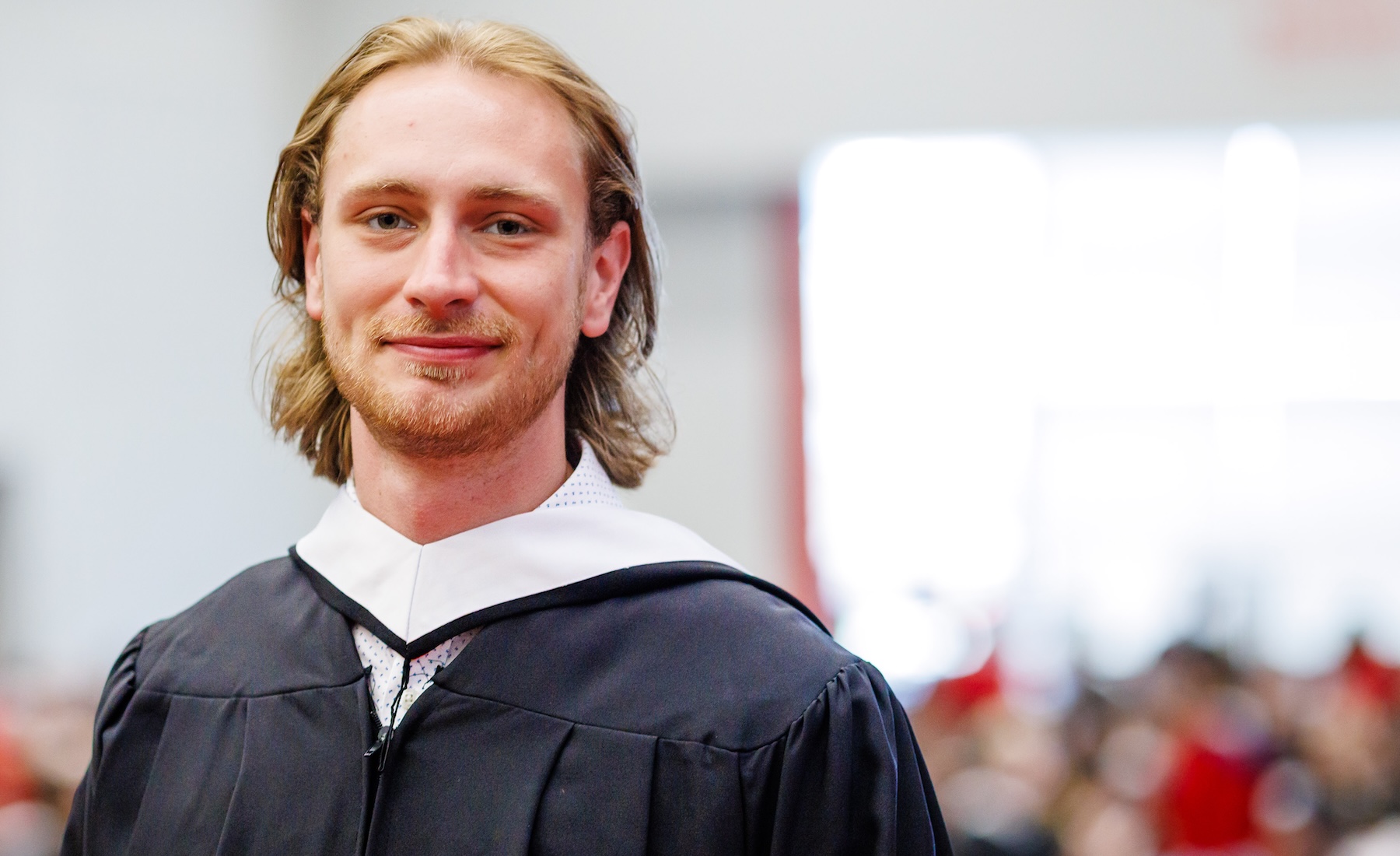 A young man in an academic robe smiles while standing in front of an audience at a university graduation ceremony. 