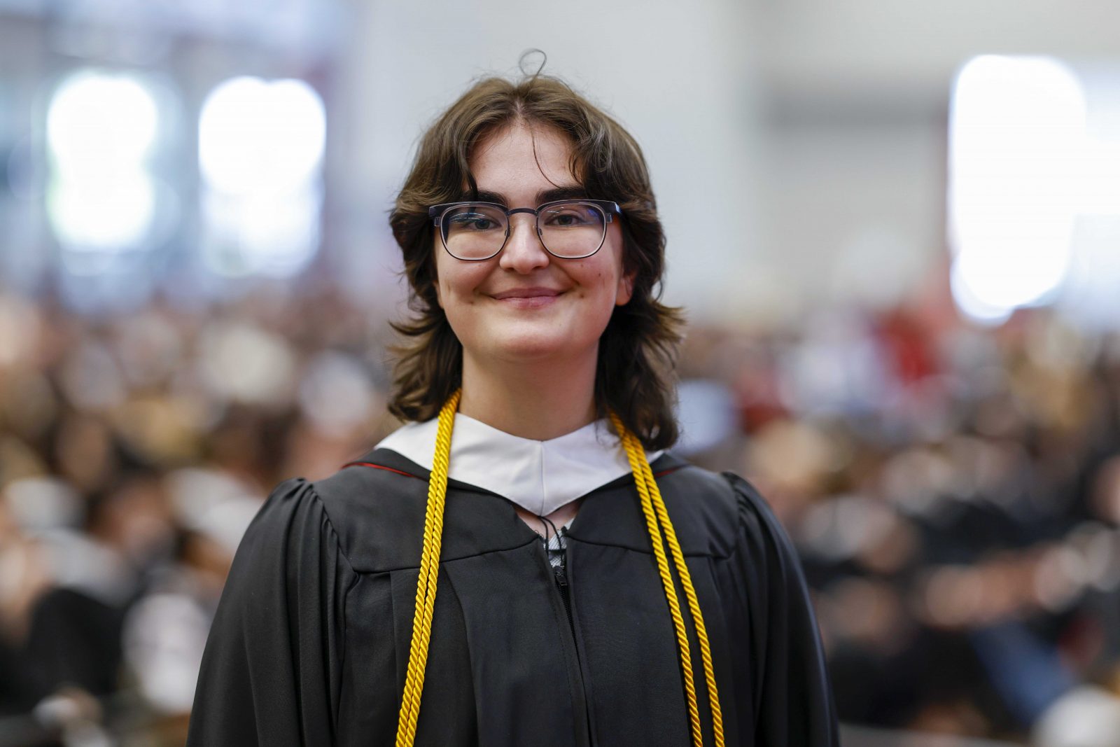 A person in an academic gown smiles while standing in a university graduation ceremony with an audience visible in the background.