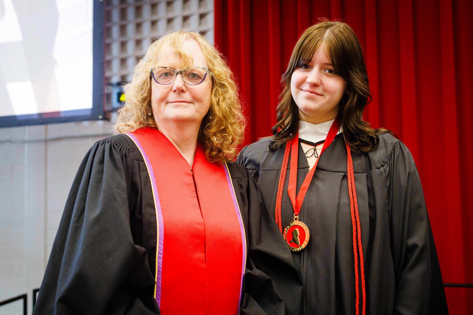 Two women stand on stage dressed in convocation regalia smiling at the camera.