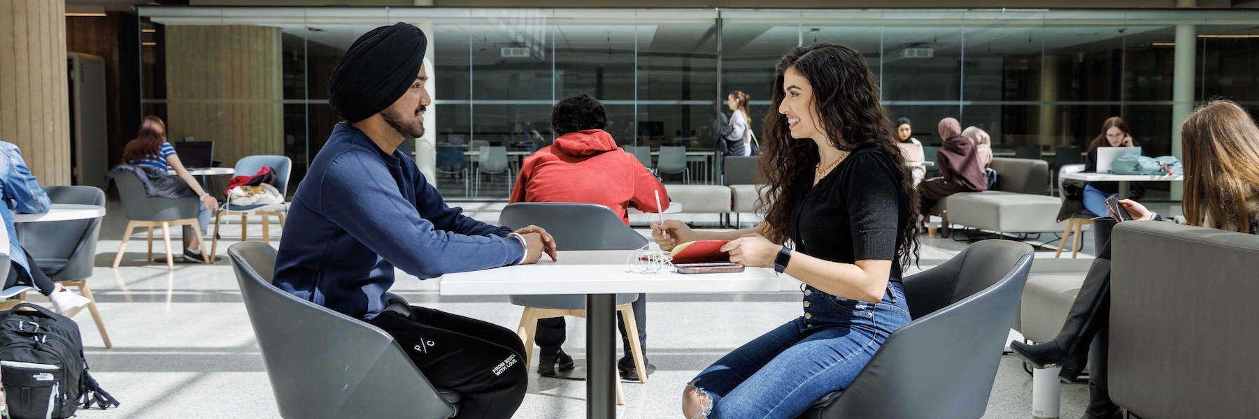 Two students sitting across from each other at a table in a brightly lit atrium.