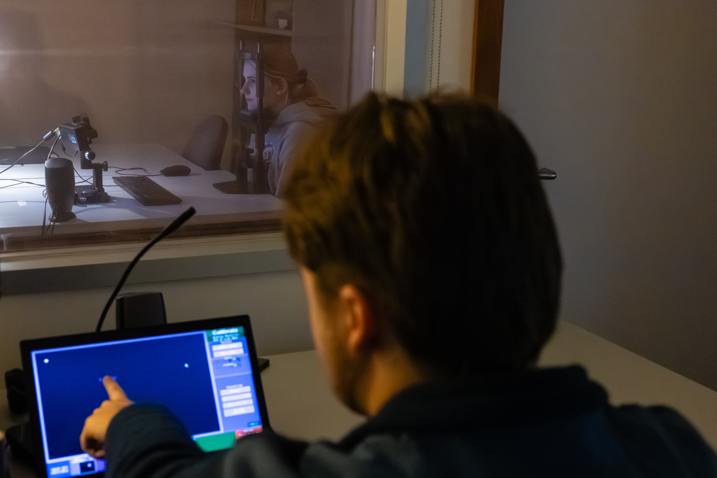 A person points at data on a laptop while looking through a lab room window where a person is sitting at a desk with their chin propped on a mechanism.
