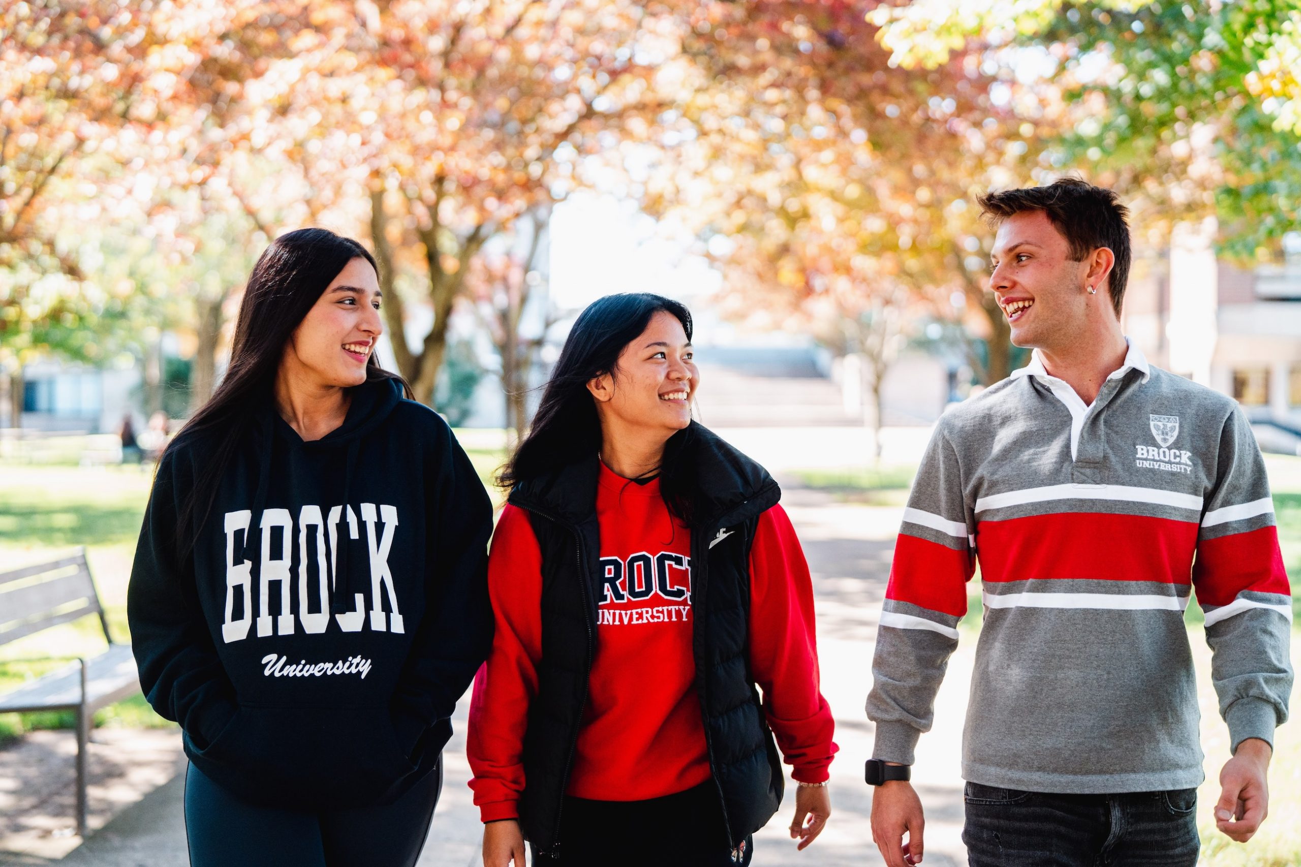 Three students in Brock University sweaters walking on campus with fall-coloured trees in the background.