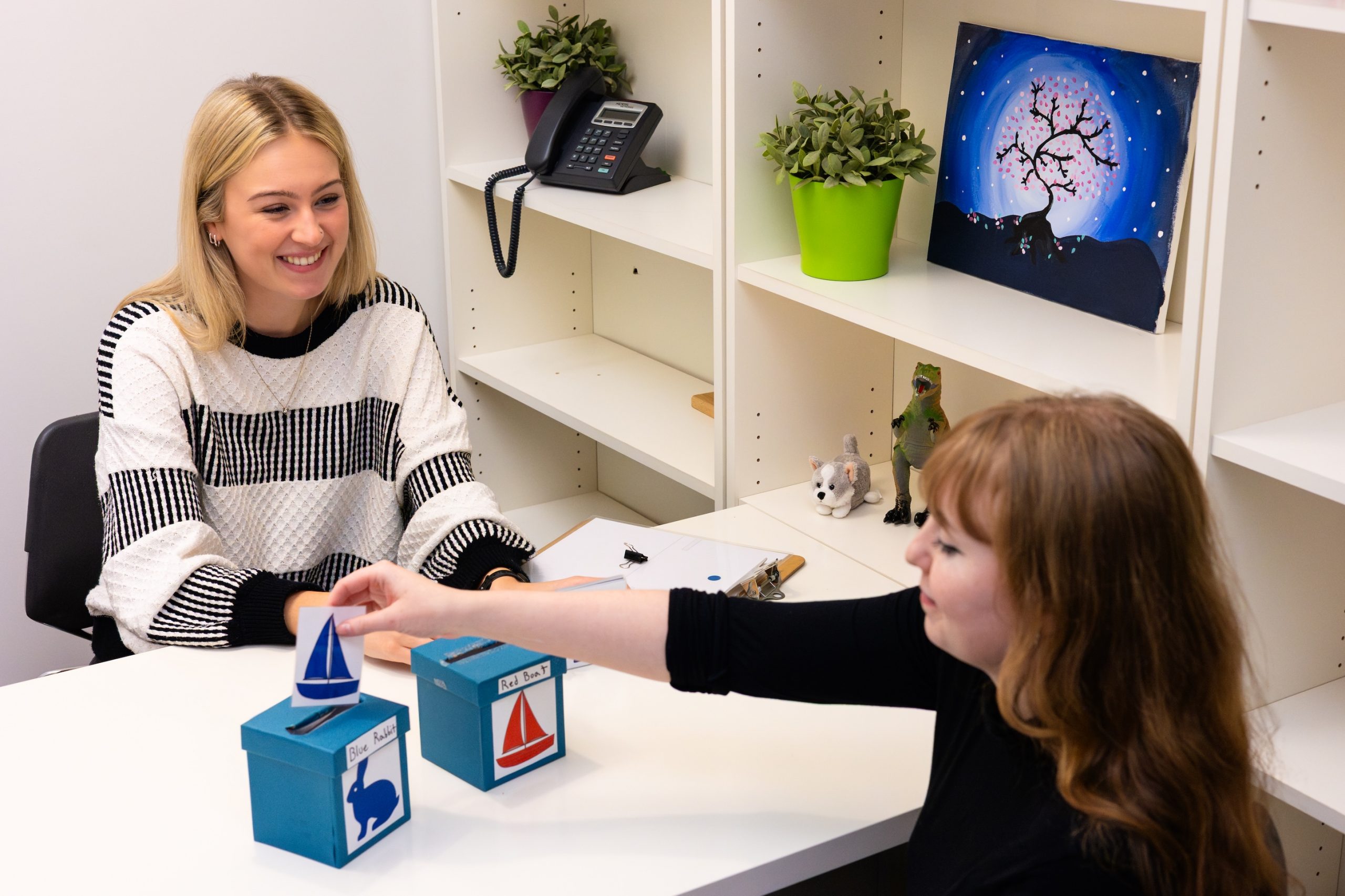 Two people sit facing each other at a table while one person inserts a card with a blue sailboat into a box with a blue rabbit illustration on it.