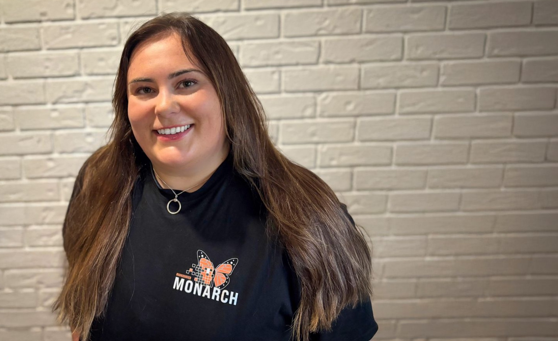 A female Brock University Psychology student, smiling while wearing a black Project Monarch T-shirt, standing against a white brick wall. 