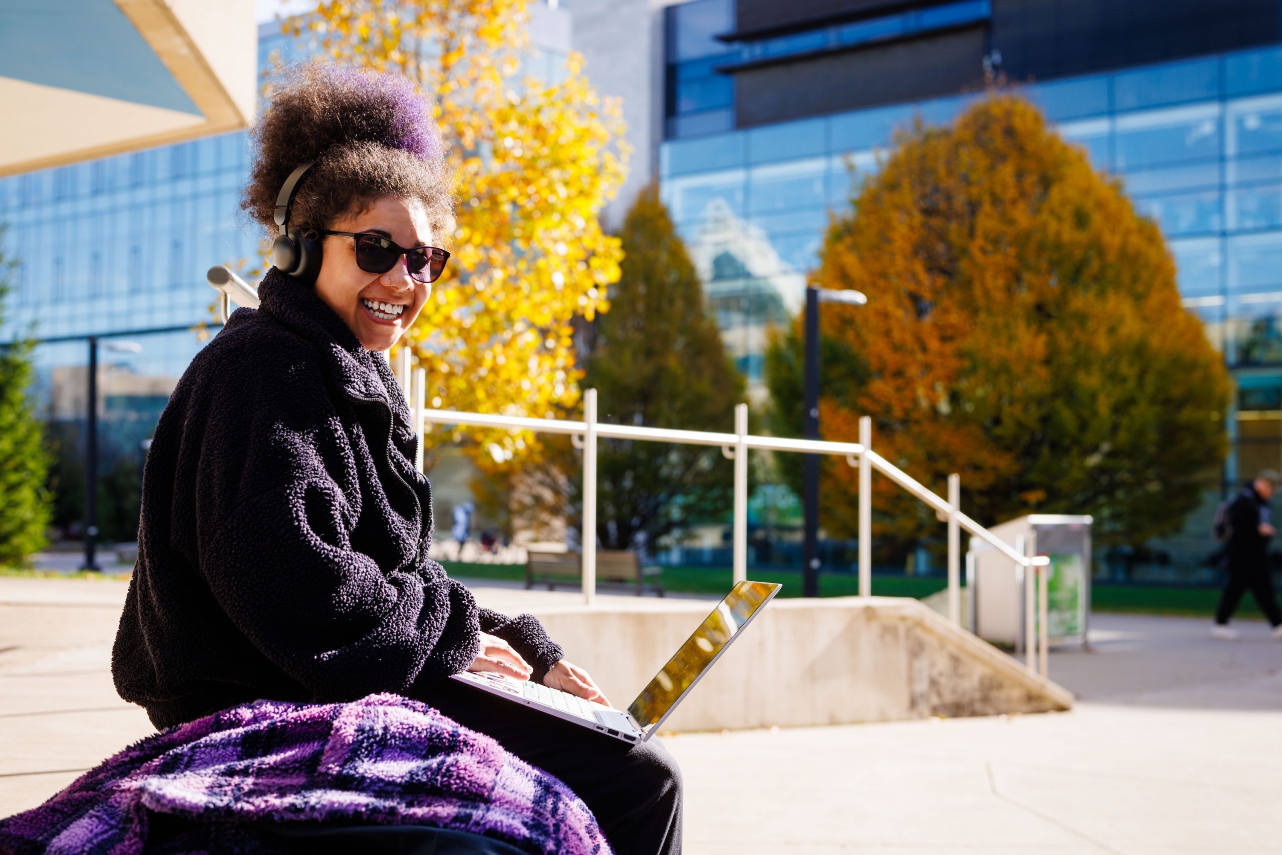 A person sitting outside on a bench with a laptop and fall-coloured trees in the background.