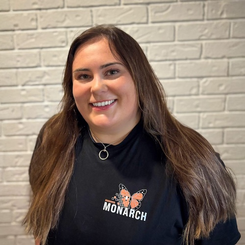 A female Brock University Psychology student, smiling while wearing a black Project Monarch T-shirt, standing against a white brick wall.