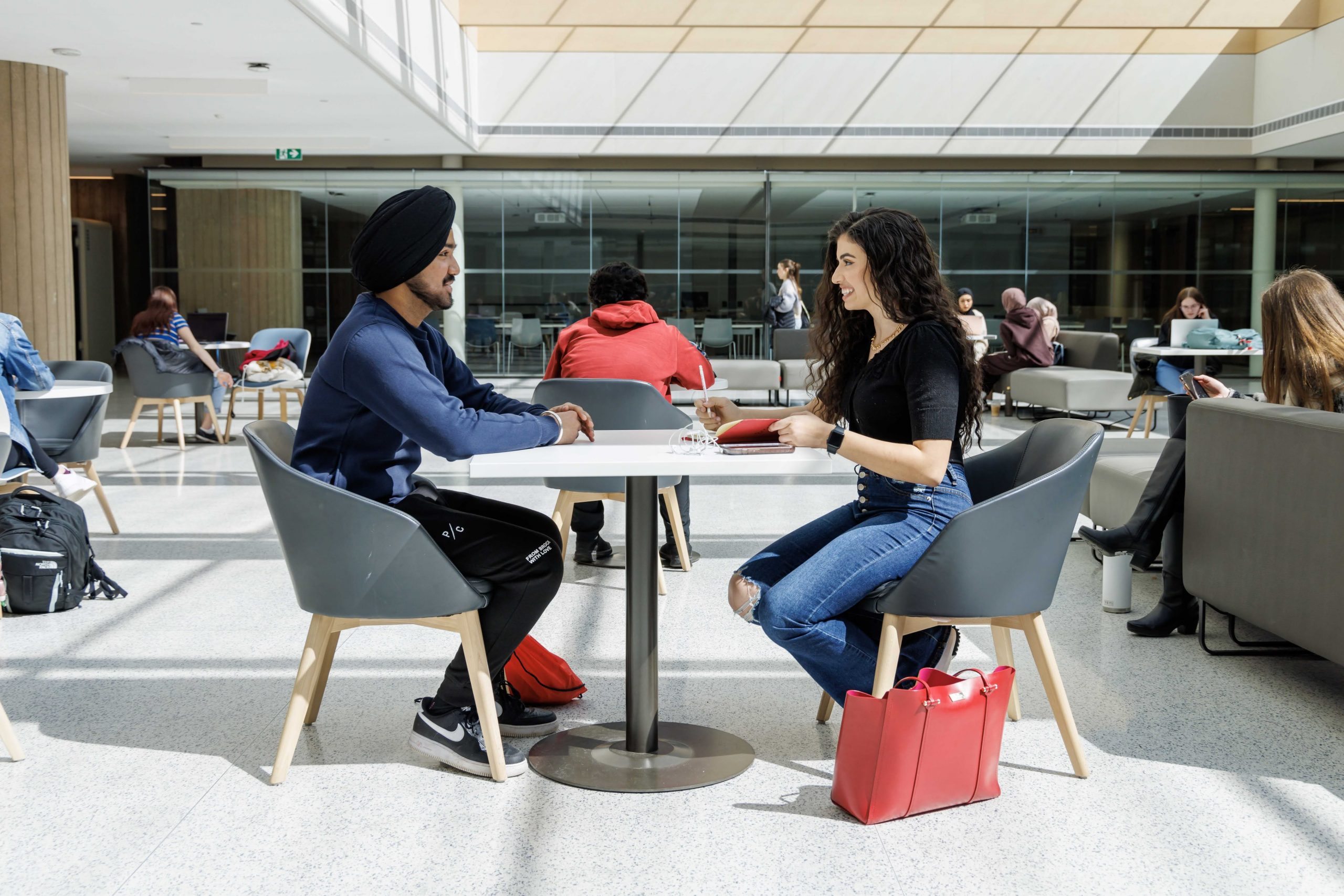 Two students sitting across from each other at a table in a brightly lit atrium.