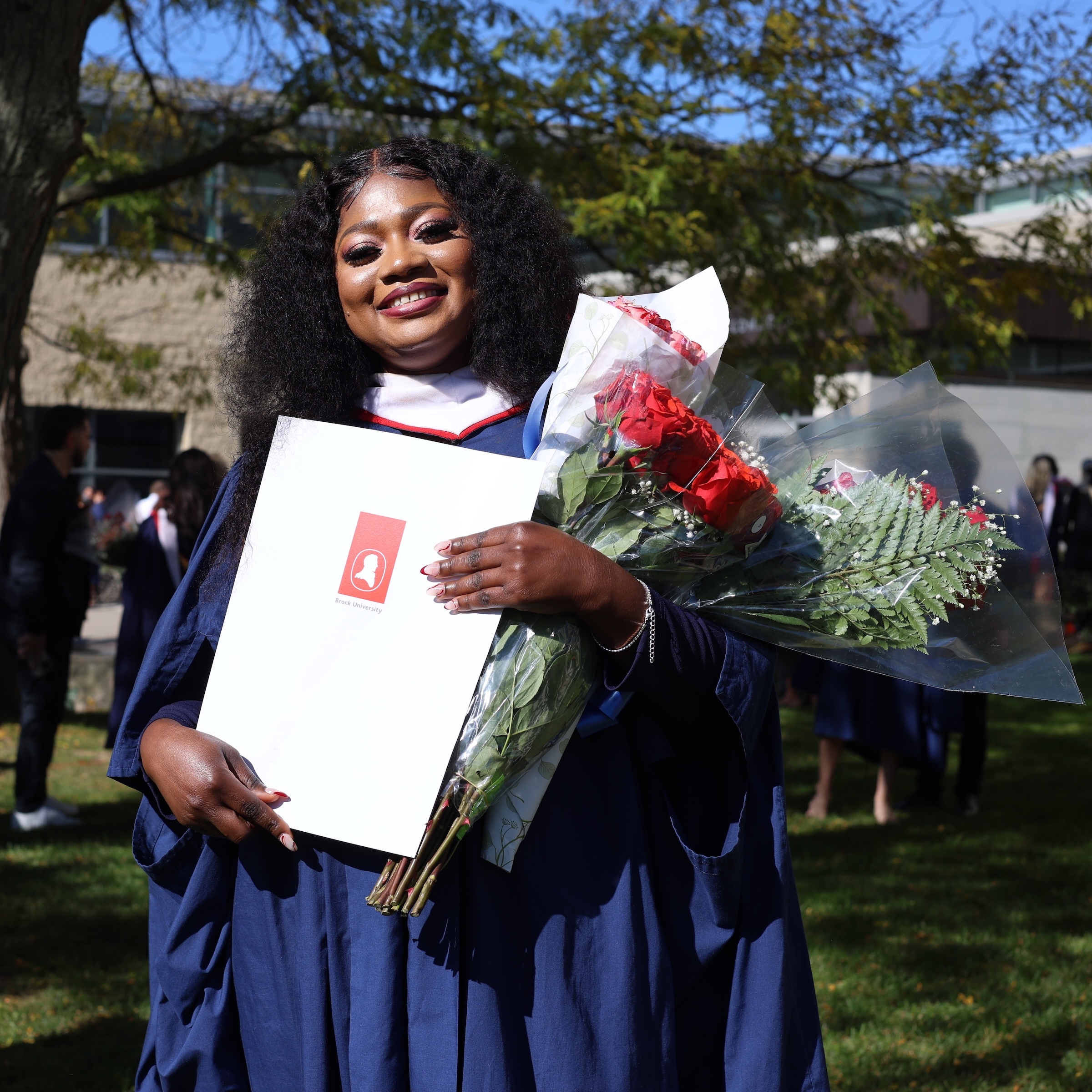 Laadi Salifu (MA ’24) stands outside on Brock University's main campus wearing a graduation gown, holding bouquets of red roses and an envelope her degree.