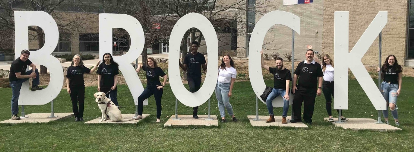 Group of ten individuals and a service dog posing with the Brock letters on Brock University's main campus.