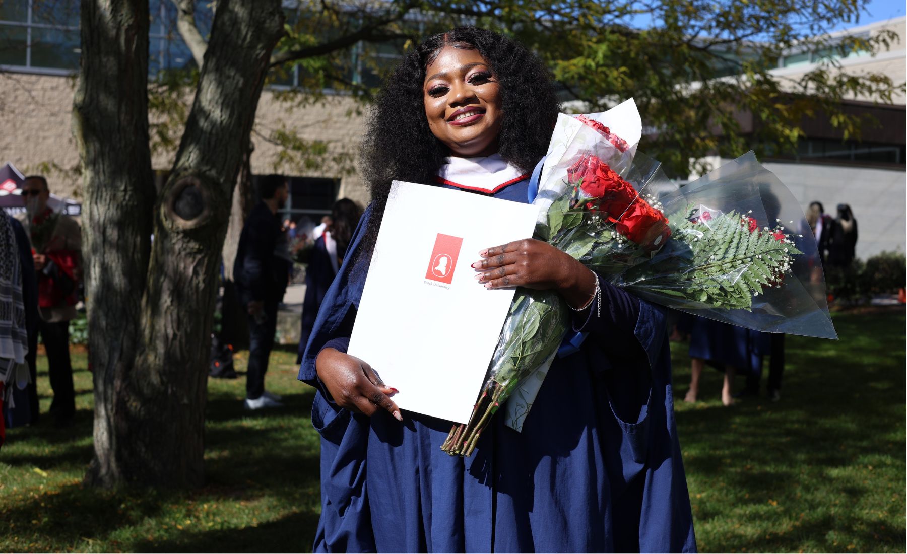 Laadi Salifu (MA ’24) stands outside on Brock University's main campus wearing a graduation gown, holding bouquets of red roses and an envelope her degree. 