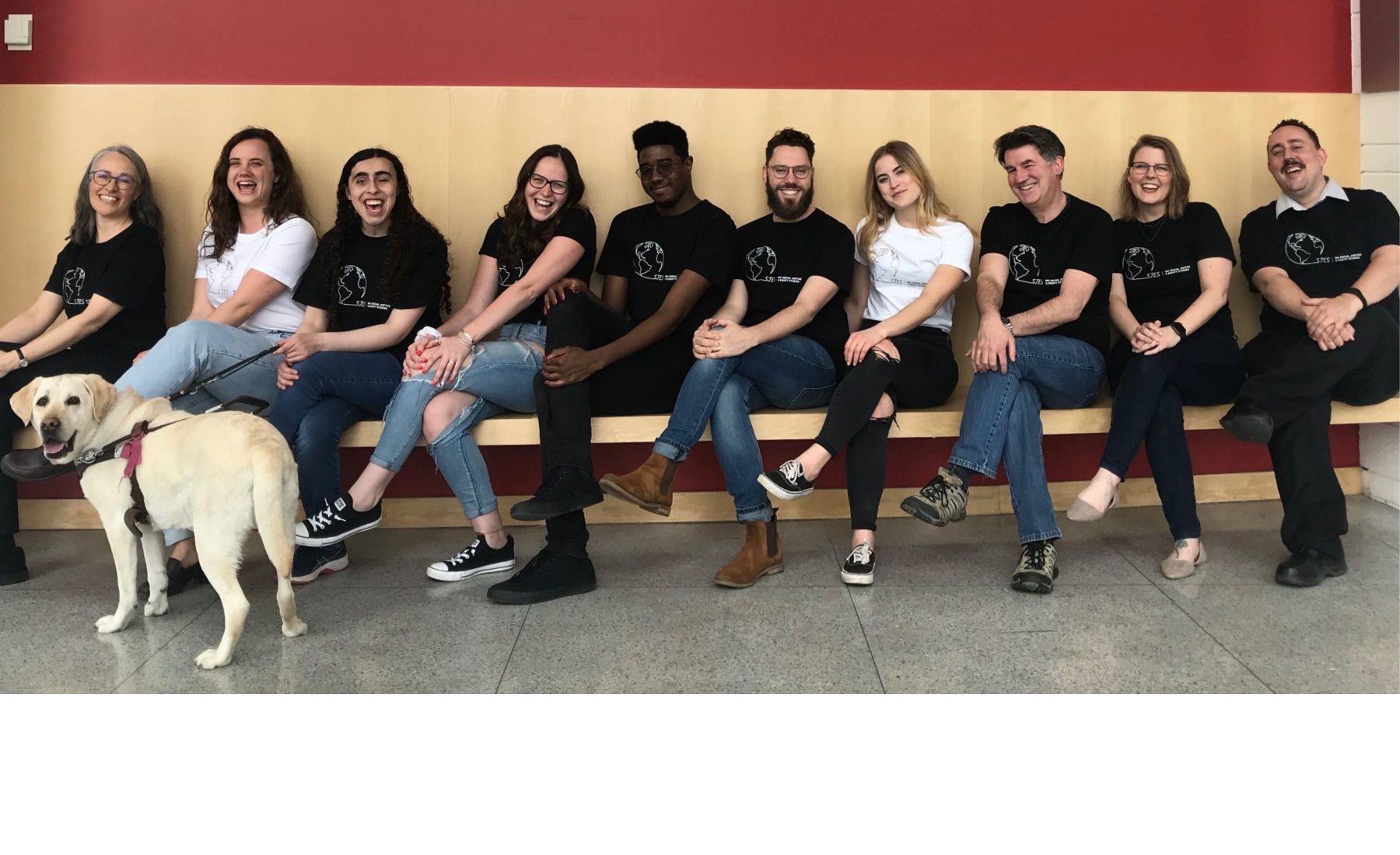 Group of students and professors sitting in a line with their legs crossed on a bench. A service dog sits at one end. 