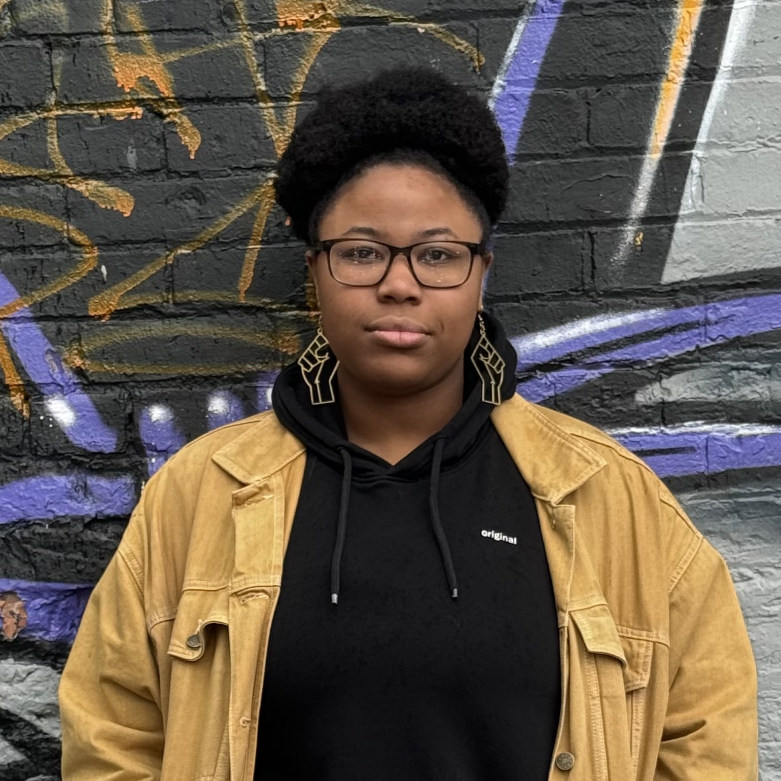 Alli Rolle standing in front of a brick wall with colourful graffiti.