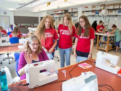 Three students wearing Brock Cares shirts stand behind a community member demonstrating how to use a sewing machine