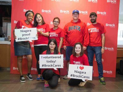 Seven students wearing red Brock shirts stand in front of a Brock banner holding signs reading: 