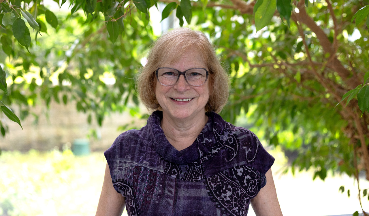 Woman wearing purple shirt and glasses stands in front of a window with trees in background.