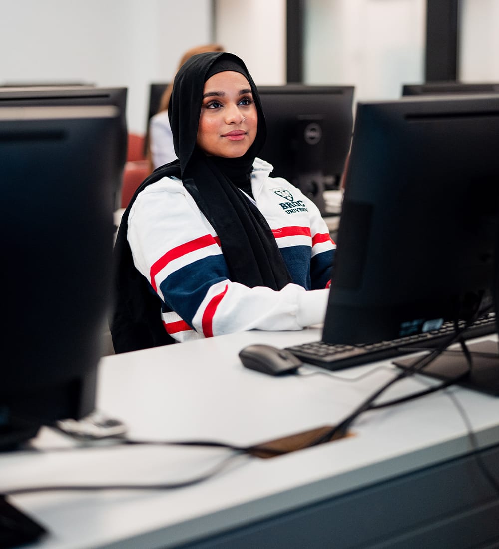 A student wearing a hijab works at a desktop computer in a university computer lab. 