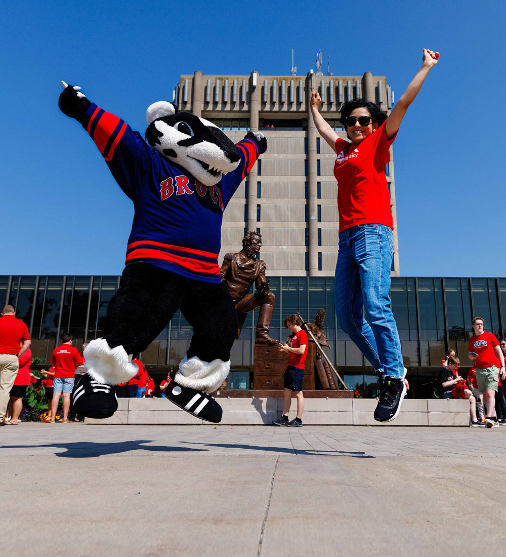 Boomer the Badger and a person in a red Brock shirt jump in the air in front of the iconic Schmon Tower. 