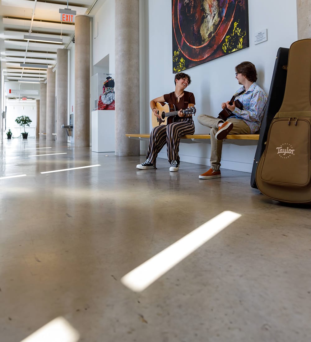 Two Brock students sit on a bench playing guitars in a sunlit hallway in the Marilyn I. Walker School of Fine and Performing Arts. 