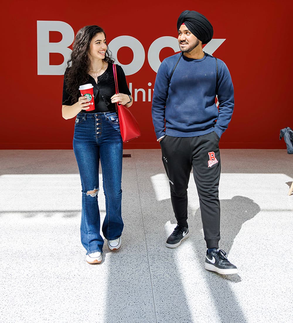 Two Brock students walking through the Rankin Family Pavilion, a large Brock logo is visible on a red wall behind them.